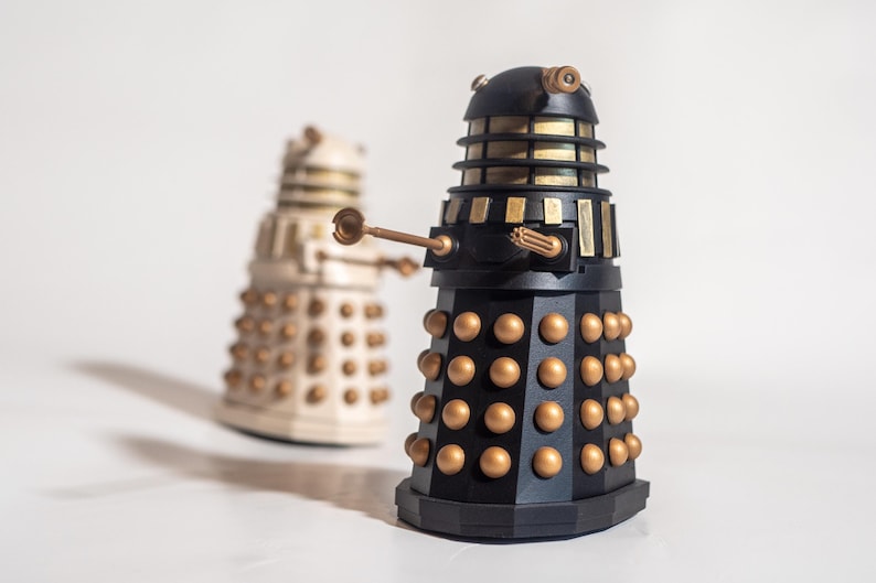 May include: Two Dalek figurines, one black and gold, the other white and gold. The black Dalek is in focus and has a raised arm with a weapon. Both Daleks have a dome-shaped head with a single eye.