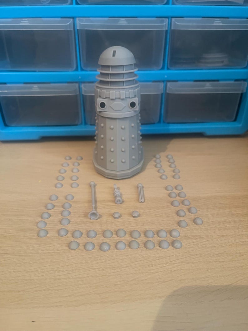 May include: A gray plastic model kit of a Dalek, a fictional robot from the British science fiction television series Doctor Who. The kit includes the main body of the Dalek, three guns, and a number of small dome-shaped pieces.