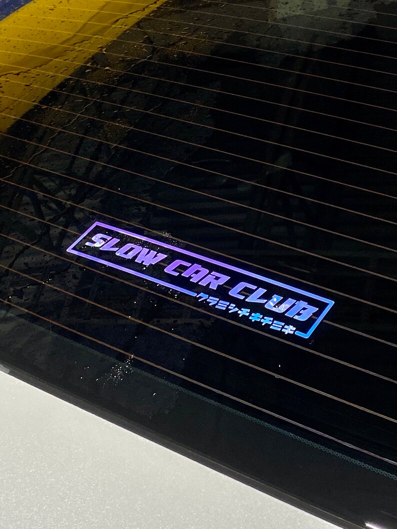 May include: A rectangular sticker applied to a dark glass surface, featuring the text SLOW CAR CLUB in a bold, stylised font with a blue outline. Below the main text, smaller Japanese characters are printed within the same blue border design.