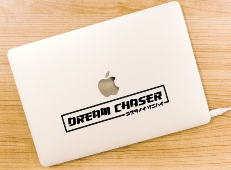 May include: A silver laptop featuring a black rectangular decal with the text DREAM CHASER and Japanese characters inside a border. The sticker is positioned below the centred Apple logo on the smooth metallic surface of the closed computer.