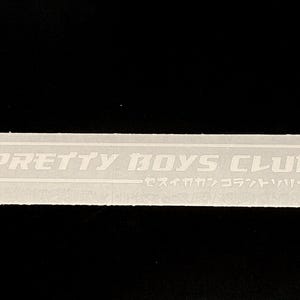 May include: A rectangular white sticker featuring the text PRETTY BOYS CLUB in a bold, stylised font enclosed within a thin white border. Below the main text, there is a line of Japanese katakana characters also enclosed in the border.
