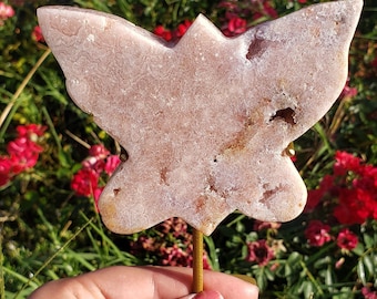 May include: A pink butterfly-shaped crystal on a gold metal stand. The crystal has a rough, textured surface and is translucent.