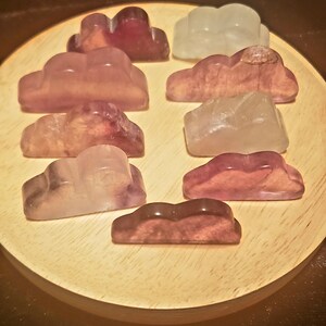 May include: Ten small, carved, cloud-shaped stones in various shades of purple, pink, and white. The stones are arranged on a wooden surface.