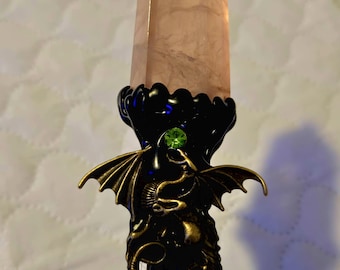 May include: A decorative wand featuring a large, light pink crystal point. The wand's handle is black with a bronze-colored dragon design and a green gem. The crystal is a light pink color.