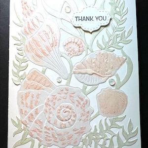 May include: A handmade greeting card with a seashell theme. The card features embossed seashells in shades of peach and white, with green seaweed accents. The words "THANK YOU" are printed on a small, white, cloud-shaped label.
