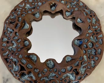Pode incluir: Um espelho redondo com uma moldura de cerâmica marrom. A moldura é decorada com um esmalte azul e branco que cria um padrão texturizado e orgânico.
