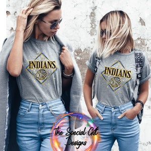 May include: Two women wearing gray t-shirts with the word "INDIANS" in gold lettering inside a diamond shape. The shirts also feature a Native American headdress graphic. They are wearing blue jeans.