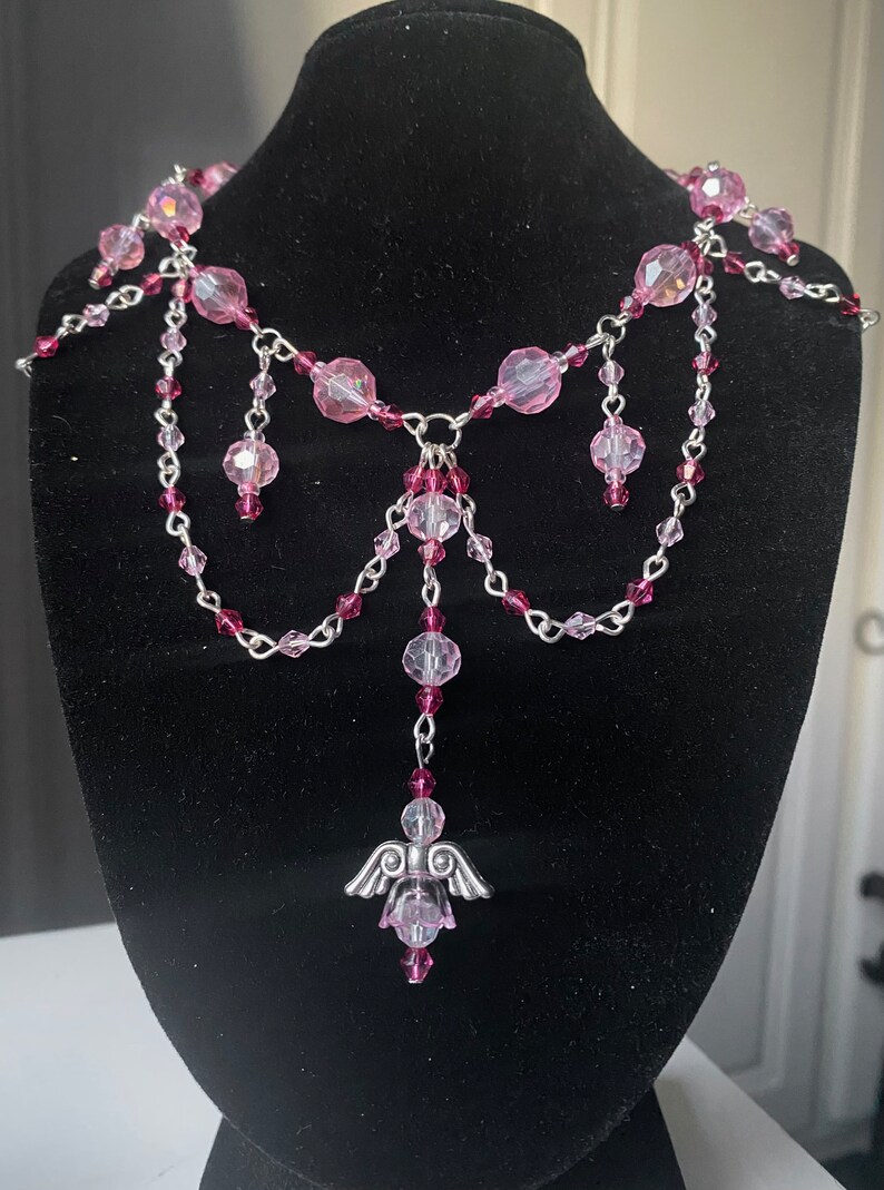 May include: A delicate pink and silver necklace with a dangling angel charm. The necklace features multiple strands of silver chain with pink crystal beads.