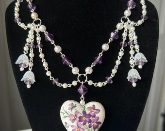 May include: A delicate necklace with a heart-shaped pendant featuring a floral design with purple flowers. The necklace is made with white pearls, silver chain, and purple crystal beads.