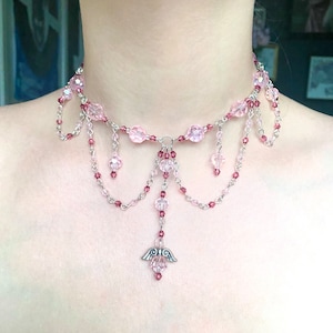 May include: A delicate pink and silver choker necklace with a dangling angel wing charm. The necklace features multiple strands of beads and chains, creating a layered and intricate design.