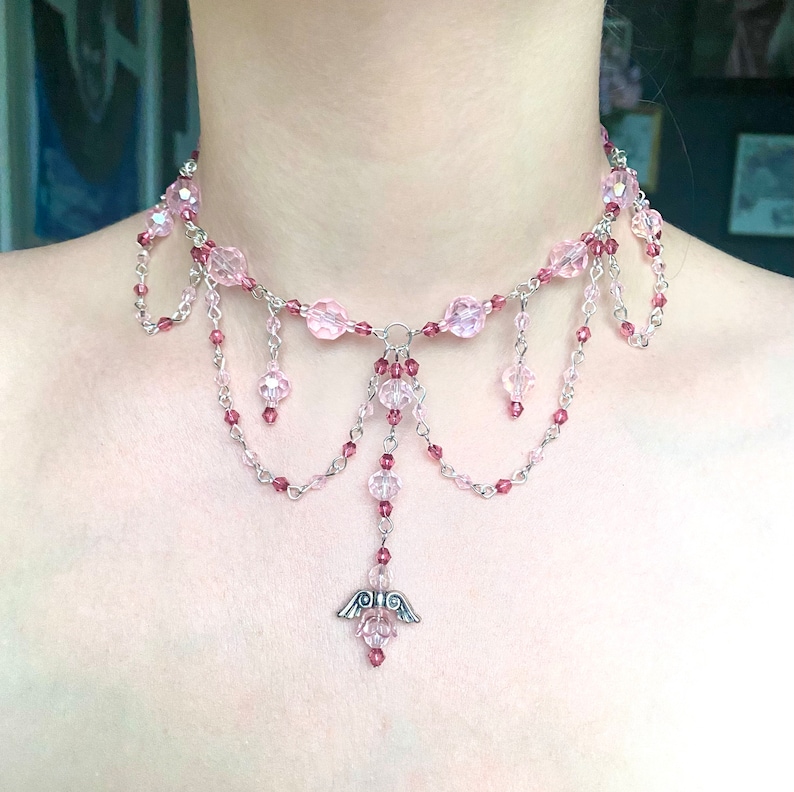 May include: A delicate pink and silver choker necklace with a dangling angel wing charm. The necklace features multiple strands of beads and chains, creating a layered and intricate design.