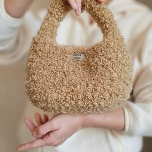 Könnte beinhalten: Eine beigefarbene, strukturierte Handtasche mit rundem Griff. Die Tasche hat ein Etikett mit der Aufschrift "ZUEVA HOME". Die Tasche besteht aus einem weichen, lockigen Material.