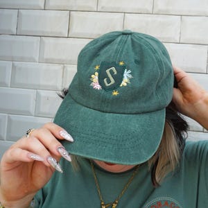 Könnte beinhalten: Eine waldgrüne Baseballkappe mit einem gestickten Wappen auf der Vorderseite. Das Wappen zeigt einen dunkelgrünen Schild mit einem stilisierten Buchstaben S in der Mitte, umgeben von kleinen gelben Sternen und bunten Blumenmotiven.