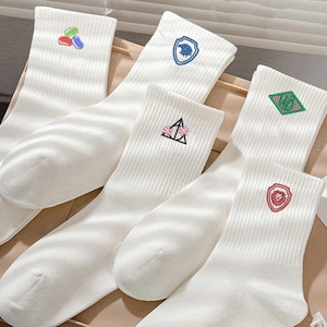 May include: Several pairs of white ribbed socks with embroidered designs. The designs include a shield with a bird, a triangle with flowers, a diamond with letters, a shield with a lion, and a cluster of colorful shapes.