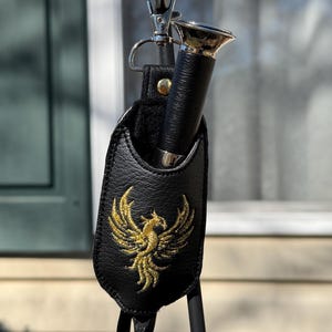 May include: A black textured leather pouch featuring a gold embroidered phoenix design on the front. The pouch contains a black tool with a silver metallic top and is attached to a silver metal swivel clasp with a small gold-toned rivet.