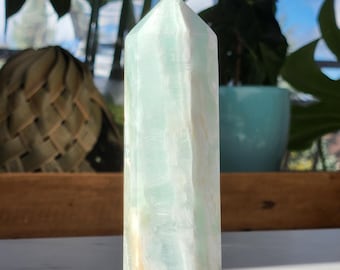 May include: A light green and white polished crystal point, also known as a wand, with a faceted shape. The crystal is translucent and has a smooth, polished surface.