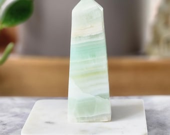 May include: A polished, rectangular crystal obelisk with a pointed top. The crystal features layers of pale green and white hues. It stands on a square, white marble base, with a blurred background of greenery and a neutral-toned wall.