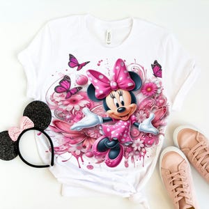 May include: White t-shirt with a pink Minnie Mouse graphic. Minnie Mouse is wearing a pink dress with white polka dots and a pink bow. The graphic is surrounded by pink flowers and butterflies.