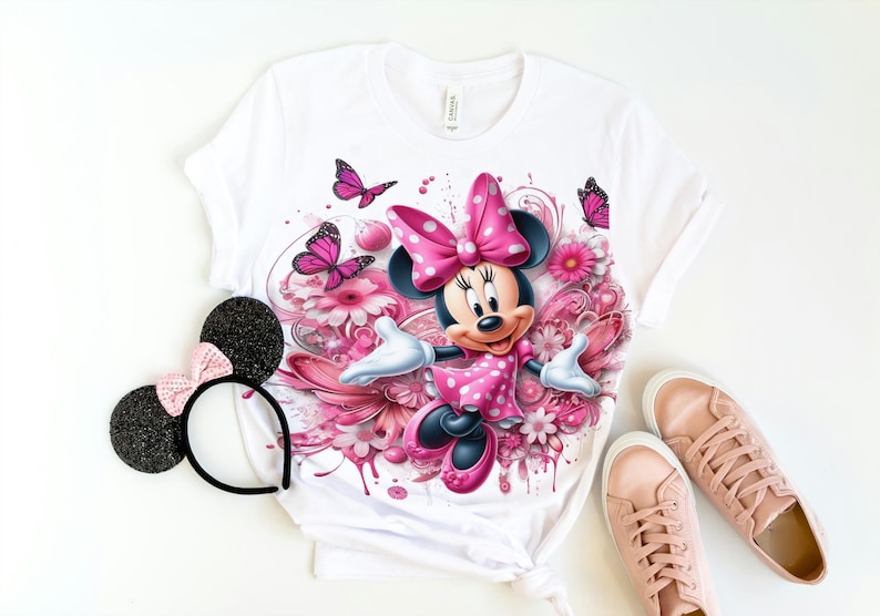 May include: White t-shirt with a pink Minnie Mouse graphic. Minnie Mouse is wearing a pink dress with white polka dots and a pink bow. The graphic is surrounded by pink flowers and butterflies.