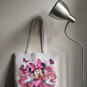 May include: White canvas tote bag with a pink and white Minnie Mouse design. Minnie Mouse is wearing a pink polka dot dress and a pink bow. The design includes pink flowers and butterflies.