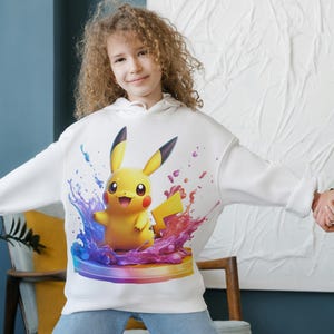 May include: White hoodie with a colorful graphic of Pikachu, a yellow electric-type Pokemon, surrounded by blue, purple, and pink paint splatters.