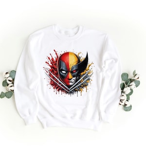 May include: White sweatshirt with a graphic print of Deadpool and Wolverine's faces, with their claws crossed, in a red, yellow, and black color scheme.