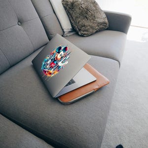 May include: A gray laptop with a colorful Mickey Mouse graphic on the lid rests on a brown leather laptop sleeve. The laptop is partially open, revealing the keyboard. The laptop and sleeve are on a gray sofa.