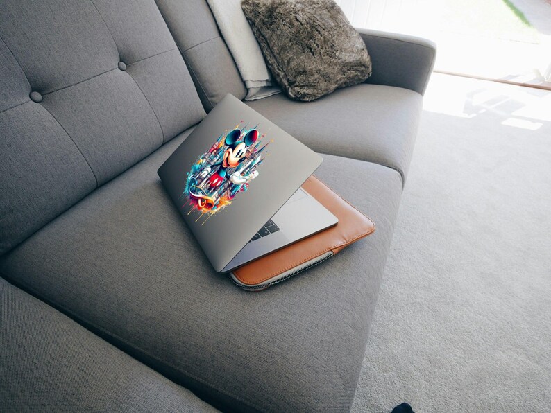 May include: A gray laptop with a colorful Mickey Mouse graphic on the lid rests on a brown leather laptop sleeve. The laptop is partially open, revealing the keyboard. The laptop and sleeve are on a gray sofa.