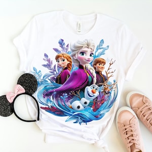 Pu&ograve; includere: Maglietta bianca con una stampa grafica colorata di Elsa, Anna e Olaf del film Frozen. I personaggi sono circondati da vortici blu e viola.