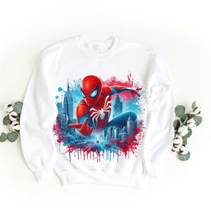 May include: White sweatshirt with a red and blue graphic print of Spiderman swinging through a cityscape. The graphic has a paint splatter effect.
