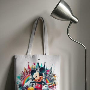May include: White tote bag featuring a colorful illustration of Mickey Mouse in front of a castle and rainbow. The design includes vibrant splashes of color and a cartoon-style aesthetic. The bag has a simple, practical design with long handles.