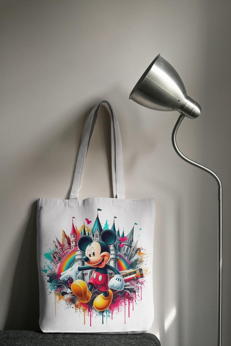 May include: White tote bag featuring a colorful illustration of Mickey Mouse in front of a castle and rainbow. The design includes vibrant splashes of color and a cartoon-style aesthetic. The bag has a simple, practical design with long handles.