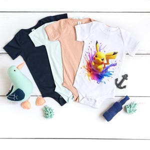 May include: Three baby onesies, one white with a colorful graphic of Pikachu, one light blue, and one peach. The onesies are laid out on a white wooden surface with a small seagull toy, a blue bottle, and two green plants.