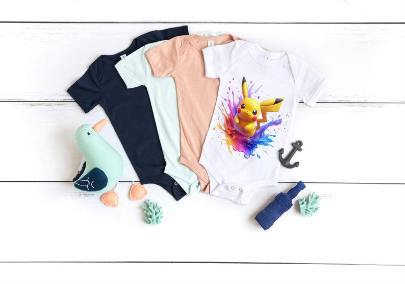 May include: Three baby onesies, one white with a colorful graphic of Pikachu, one light blue, and one peach. The onesies are laid out on a white wooden surface with a small seagull toy, a blue bottle, and two green plants.
