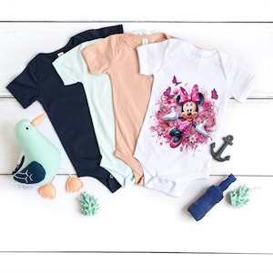 May include: Three baby onesies, one white with a pink Minnie Mouse graphic, one light blue, and one peach. The onesies are laid out on a white wooden surface.