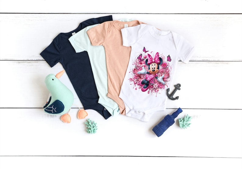 May include: Three baby onesies, one white with a pink Minnie Mouse graphic, one light blue, and one peach. The onesies are laid out on a white wooden surface.