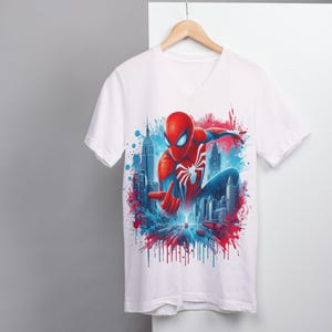 May include: White t-shirt with a colorful graphic print of Spider-Man in a superhero pose, with a cityscape background and red and blue paint splatters.