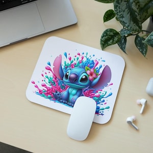 May include: A blue and white cartoon character with large ears and a flower in its hair, surrounded by pink, blue, and green paint splatters. The character is on a white mouse pad.