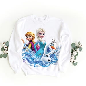 Pu&ograve; includere: Felpa bianca con una stampa colorata di Elsa, Anna e Olaf del film Frozen. I personaggi sono circondati da onde d'acqua blu.