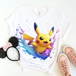 May include: White t-shirt with a colorful graphic of Pikachu, a yellow electric-type Pokemon, emerging from a splash of paint.