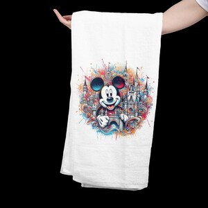May include: White hand towel featuring a colorful graphic of Mickey Mouse in front of a castle. The design includes vibrant splashes of red, blue, yellow, and green. The towel is held up against a black background.