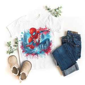 May include: White t-shirt with a colorful graphic of Spiderman swinging through the city. The graphic has red, blue, and white paint splatters.
