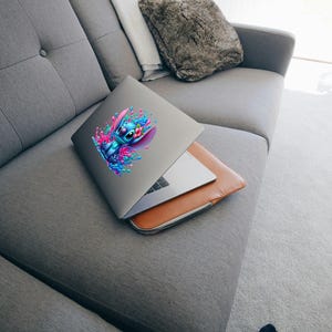 May include: A silver laptop with a blue and pink cartoon character sticker on the lid. The laptop is closed and sitting on a gray couch.