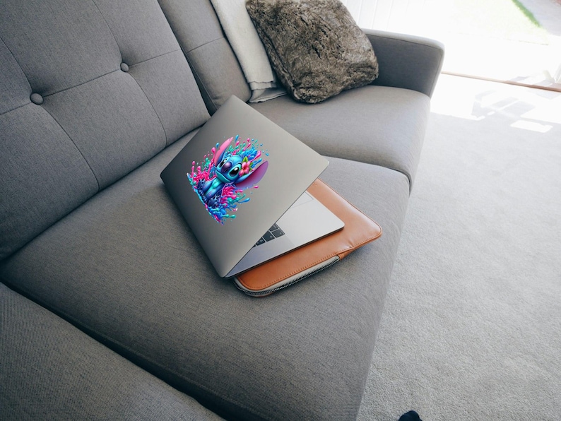 May include: A silver laptop with a blue and pink cartoon character sticker on the lid. The laptop is closed and sitting on a gray couch.