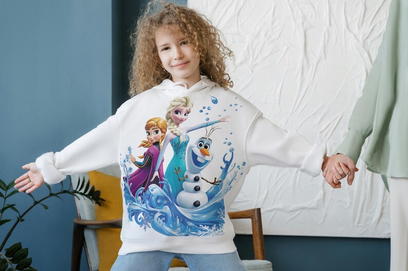 Pu&ograve; includere: Felpa con cappuccio bianca con una stampa grafica colorata dei personaggi Elsa, Anna e Olaf del film Frozen. I personaggi sono raffigurati in una scena invernale con fiocchi di neve e ghiaccio.