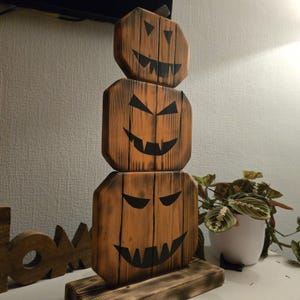 Può includere: Una pila verticale di tre forme di zucca in legno montate su una base rettangolare. Ogni zucca presenta una finitura in legno bruciato con venature marrone scuro e volti di zucca dipinti in nero con occhi triangolari e bocche dentate.