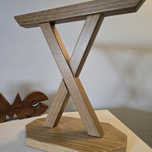 Könnte beinhalten: Ein dekorativer Holzständer mit hellbraunem Finish. Der Ständer hat eine rechteckige Oberseite, die von zwei gekreuzten Balken getragen wird und auf einer vielseitigen Basis ruht. Die Holzmaserung ist sichtbar.