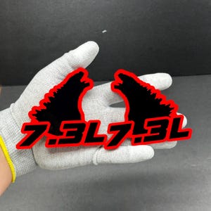 以下が含まれることがあります： 黒と赤の7.3Lステッカー2枚。各ステッカーには、太字の赤いフォントで「7.3L」というテキストが付いたモンスターのシルエットが描かれています。ステッカーは灰色の背景に表示されています。