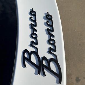 May include: Black "Bronco" lettering, in a stylized font, is affixed to a white surface. The letters cast a shadow, suggesting a raised profile. The image is a close-up of a vehicle detail, likely a decal or emblem.