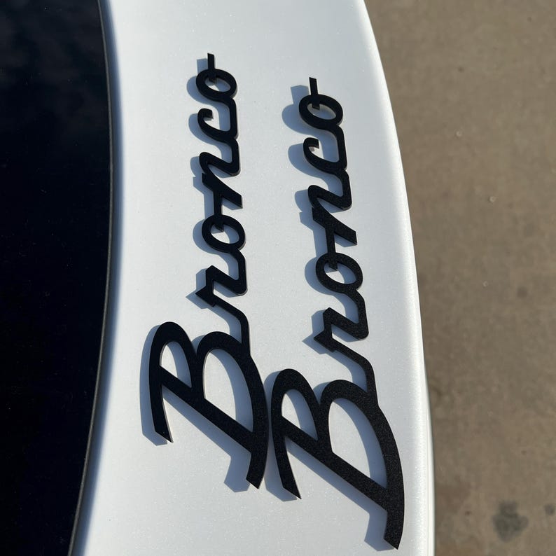 May include: Black "Bronco" lettering, in a stylized font, is affixed to a white surface. The letters cast a shadow, suggesting a raised profile. The image is a close-up of a vehicle detail, likely a decal or emblem.